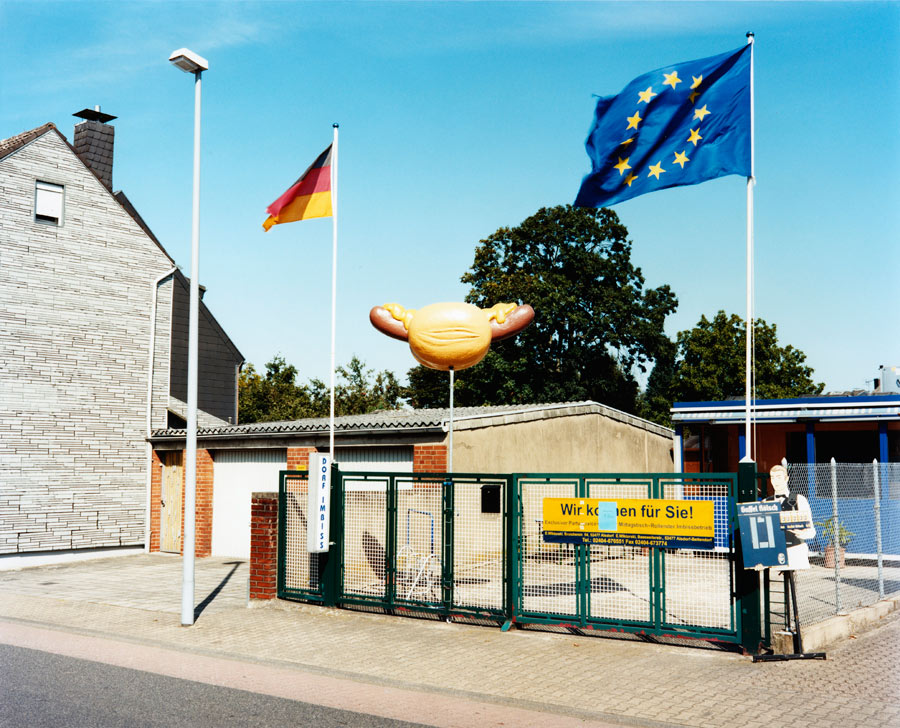 ein großes brötchen mit wurst und senf wirbt für einen dorfimbiss mit deutschland- und europaflagge. // a large roll with sausage and mustard advertises a village snack bar with a hoisted german and european flag.
