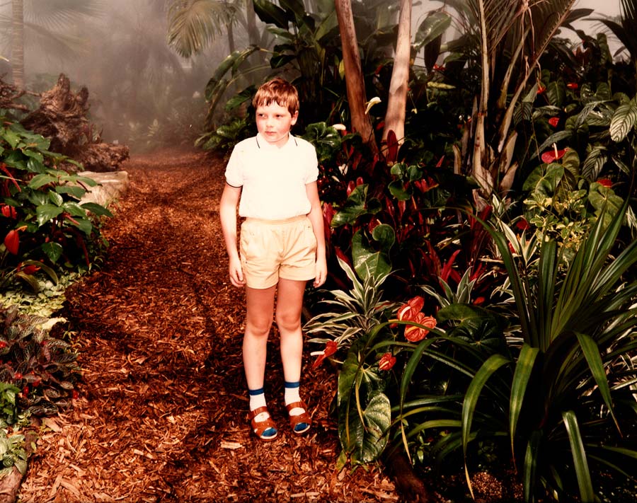 im tropenhaus; ein kleiner junge mit kurzer hose, socken und sandalen wird von seinen eltern photographiert.
