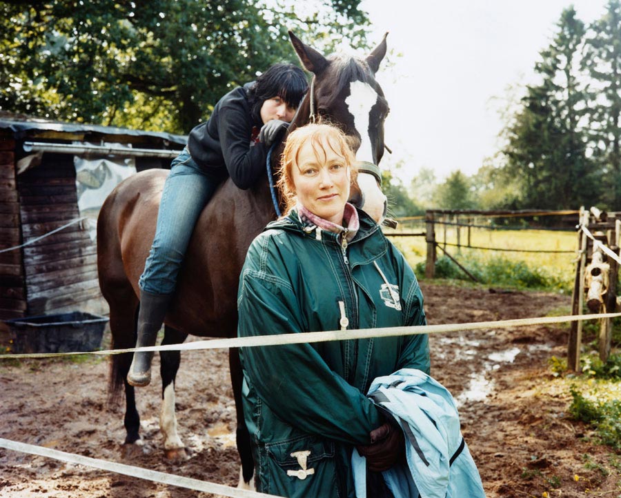 eine mutter mit ihrer tochter auf der pferdekoppel, die auf ihrem pferd sitzt und den hals des pferdes umarmt. // a mother with her daughter in the paddock, sitting on her horse and hugging the horse's neck.